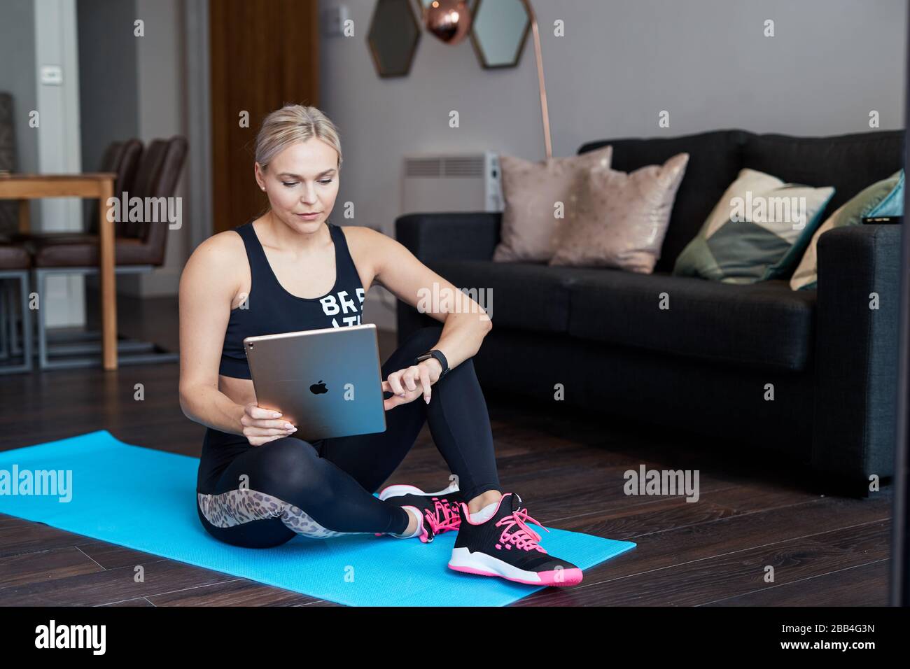 Eine Single Blond-Frau überprüft ihr Tablett, während sie zu Hause auf einer Matte im Wohnzimmer ihrer Wohnung trainiert Stockfoto