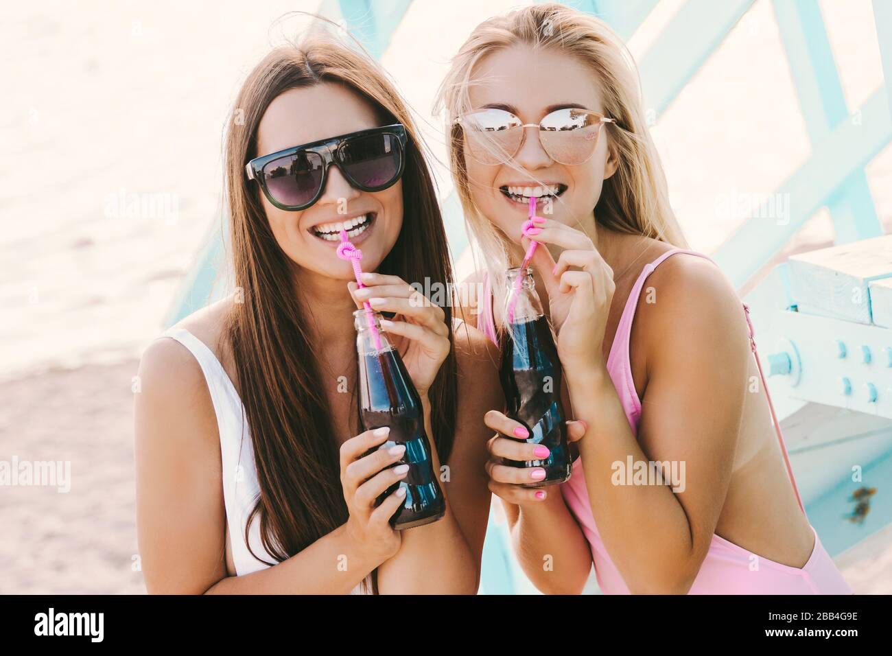 Fröhliche junge Mädchen in stylischen Badeanzügen und Sonnenbrillen, die Limonade trinken und an der Sommerküste lachen. Schöne fröhliche Frauen Freunde, die Spaß haben Stockfoto