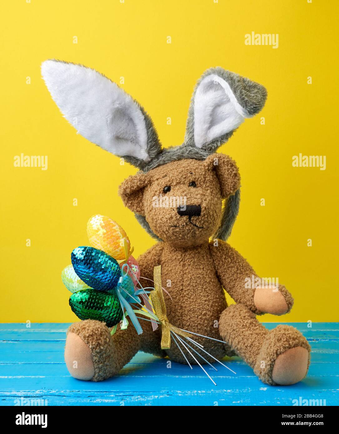 Ein süßer, brauner Teddybär aus der Vintage, der bunte Ostereier hält und eine Kaninchenmaske mit langen Ohren am Kopf trägt, lustige Ferienkarte Stockfoto