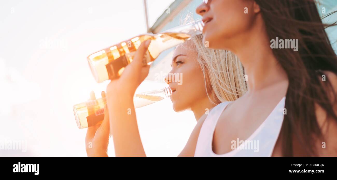 In stilvollen Badeanzügen, die Bier trinken und Spaß am sonnigen Strand haben, schließen sich zwei attraktive, fröhliche Mädchen an. Schöne junge Frauen posieren mit B. Stockfoto