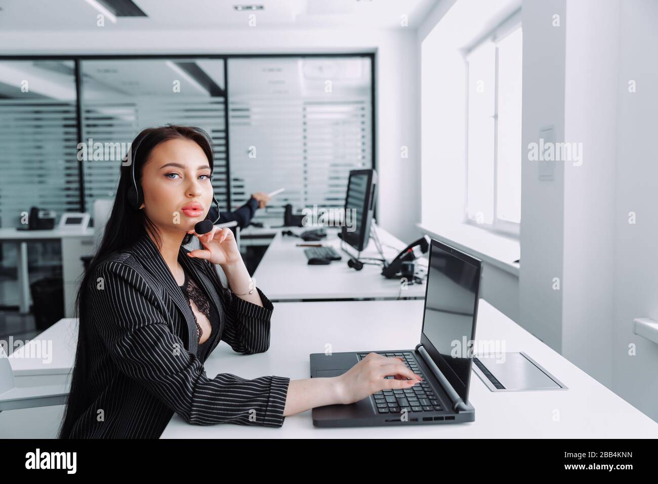 Seitenansicht einer schönen jungen Geschäftsfrau in einem Headset mit Laptop während der Arbeit im Büro. Das Mädchen sieht sich die Kamera an Stockfoto