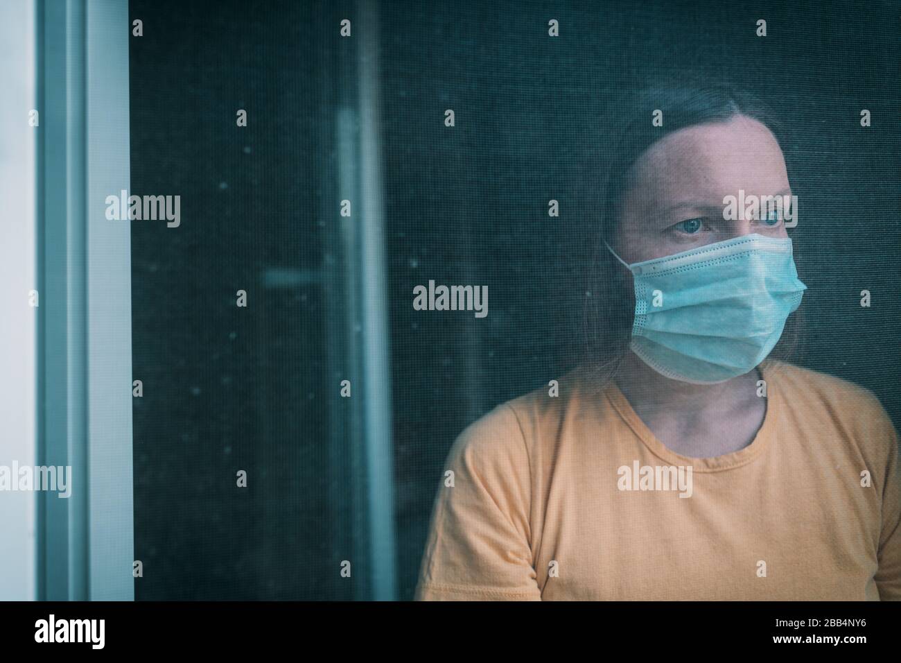 Frau in Selbstisolation beim Virusausbruch, die durch Fenster schaut. Besorgte weibliche Person mit chirurgischer Schutzmaske und selektivem Fokus Stockfoto