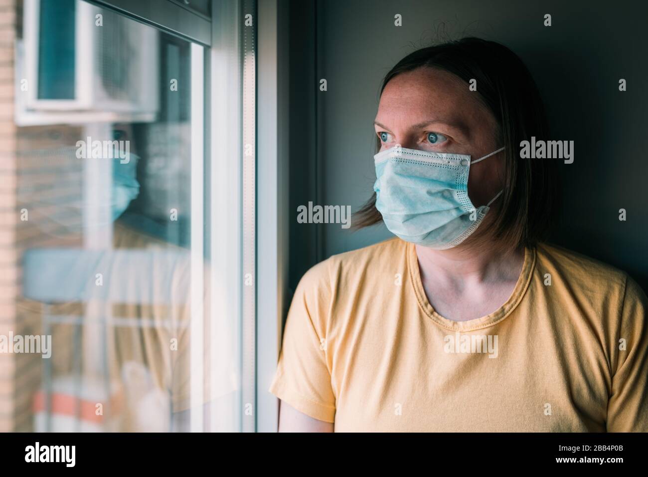 Frau in Selbstisolation beim Virusausbruch, die durch Fenster schaut. Besorgte weibliche Person mit chirurgischer Schutzmaske und selektivem Fokus Stockfoto