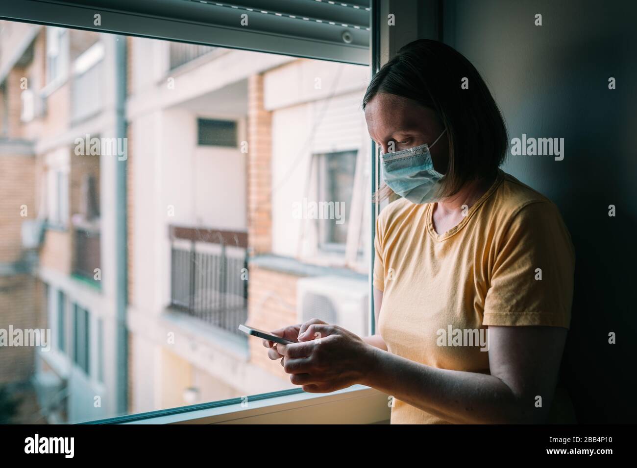 Frau in Selbstisolation bei Virusausbruch mit Handy. Besorgte weibliche Person mit chirurgischer Schutzmaske, Smartphone halten, selektiv f. Stockfoto