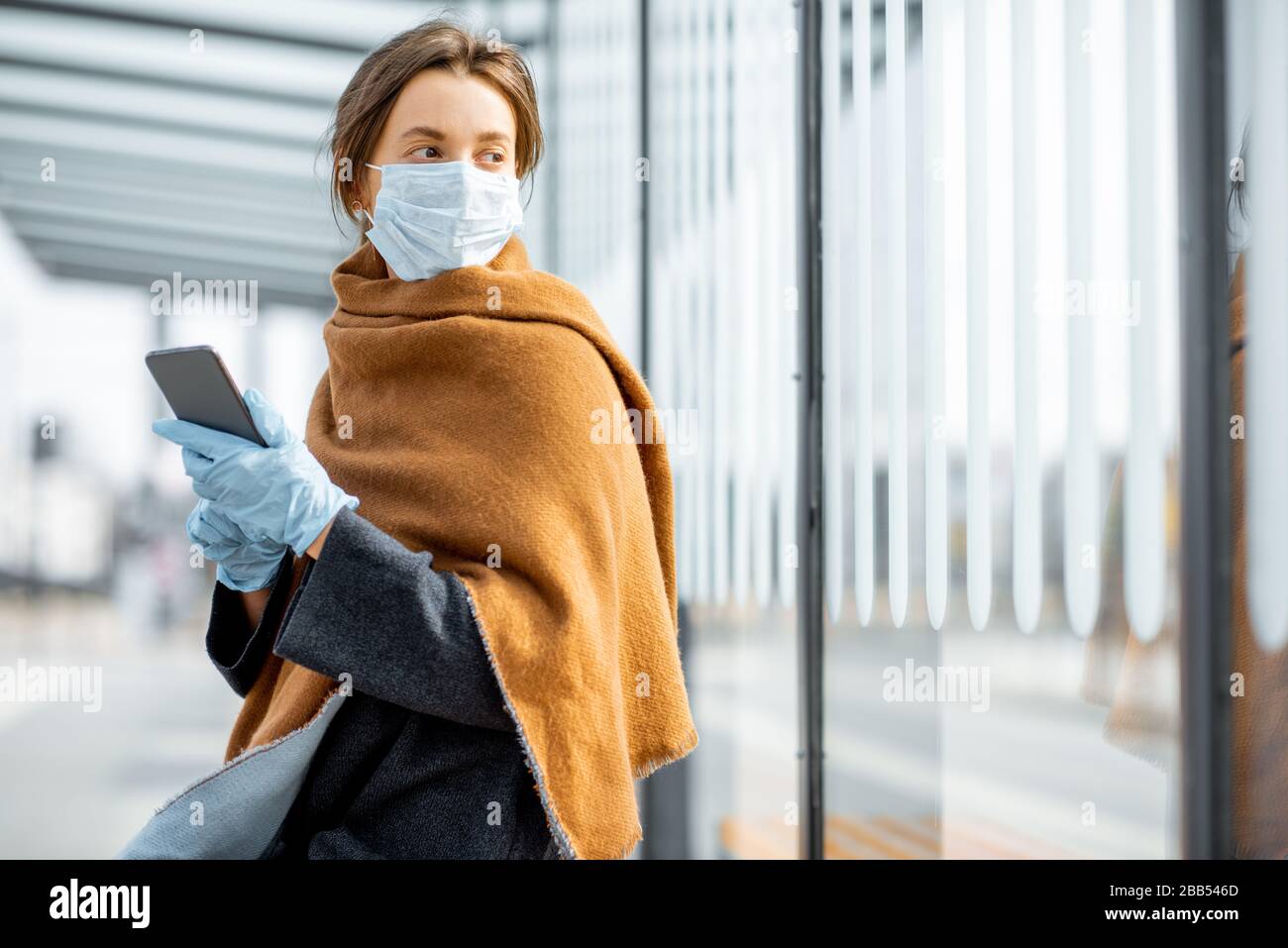 Junge Frau in Gesichtsmaske und medizinische Handschuhe mit einem Smartphone am öffentlichen Verkehrshalt während einer Epidemie. Konzept der sozialen Distanz und Online-Kommunikation Stockfoto