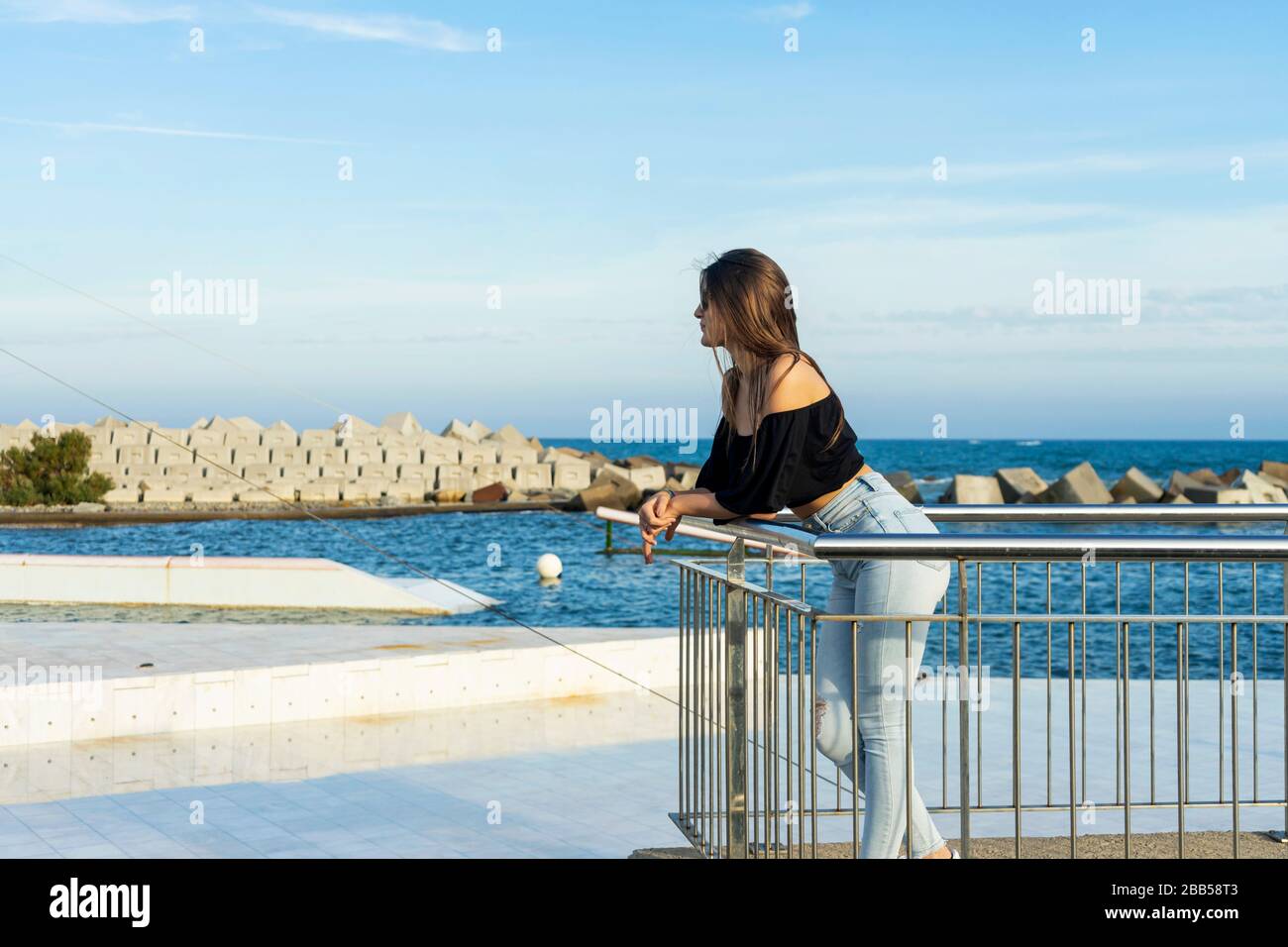 Eine junge schöne Frau mit langen Haaren, die sich am Zaun der Promenade neigt und bei Tageslicht aufdringlich beiseite blickt Stockfoto