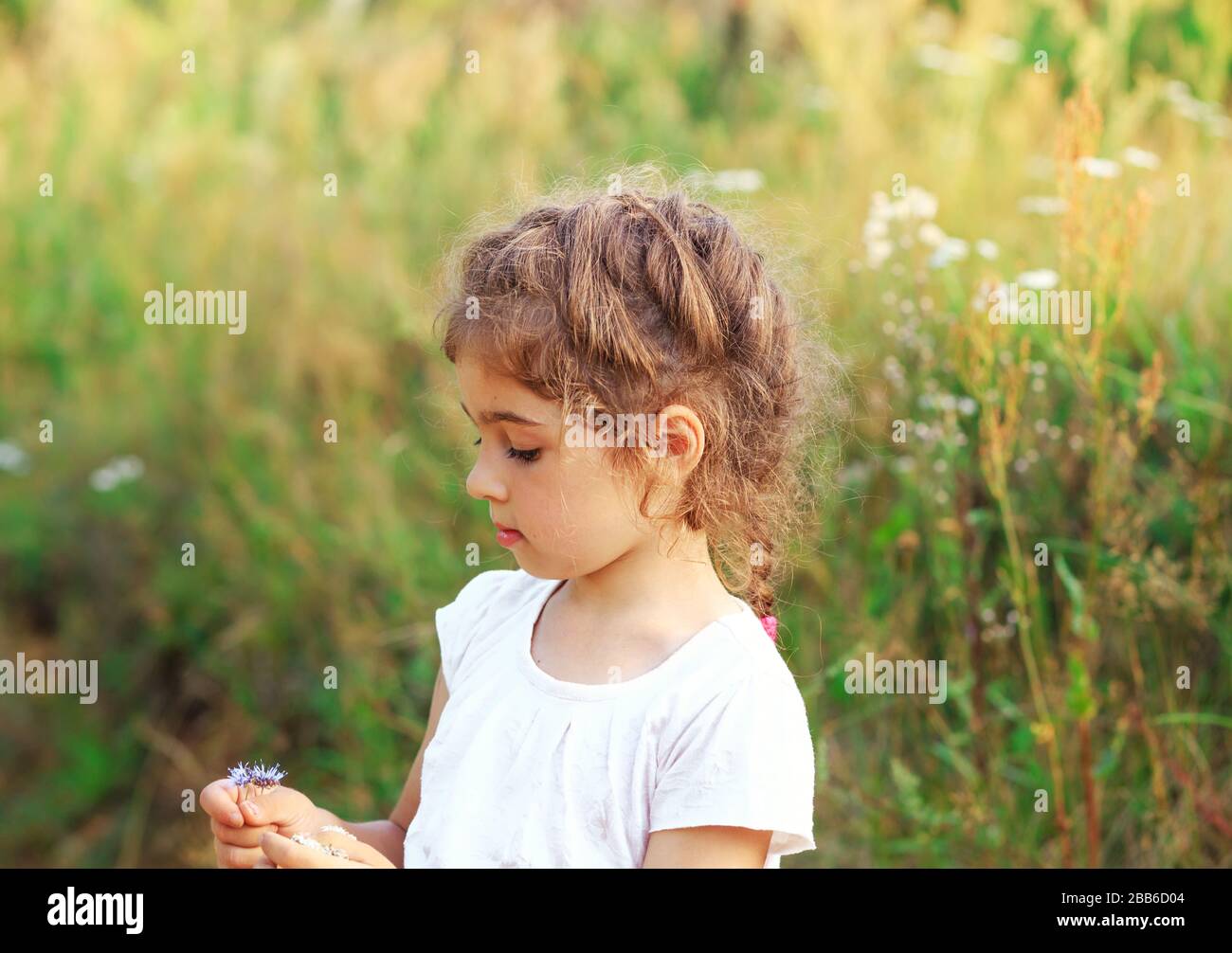 Schönes kleines Mädchen, das mit Blumen im sonnigen Frühlingspark spielt. Fröhliches süßes Kind, das bei Sonnenuntergang Spaß im Freien hat. Stockfoto