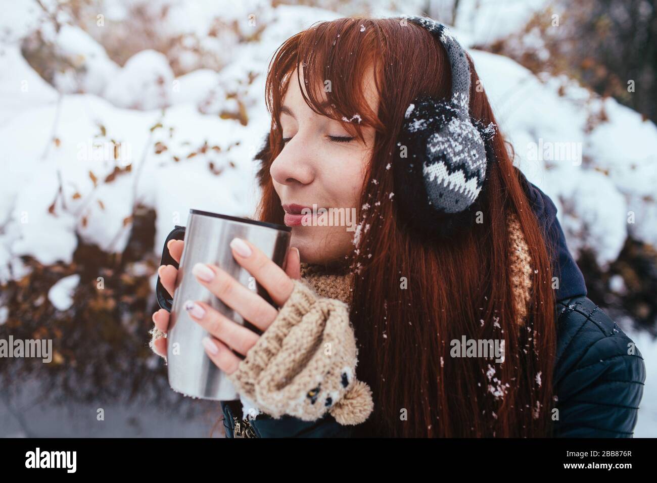 Winter. Frau mit roten Haaren, die Ohrmuscheln trägt. Mädchen trinkt heißen Tee oder Kaffee mit eiserner isolierter Tasse. Stockfoto