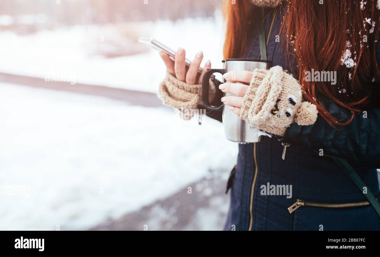 Winter. Frau mit roten Haaren, die Handschuhe trägt. Mädchen trinkt heißen Tee oder Kaffee mit eiserner isolierter Tasse. Es verwendet ein Telefon oder Smartphone. Spielen, Klo King Stockfoto