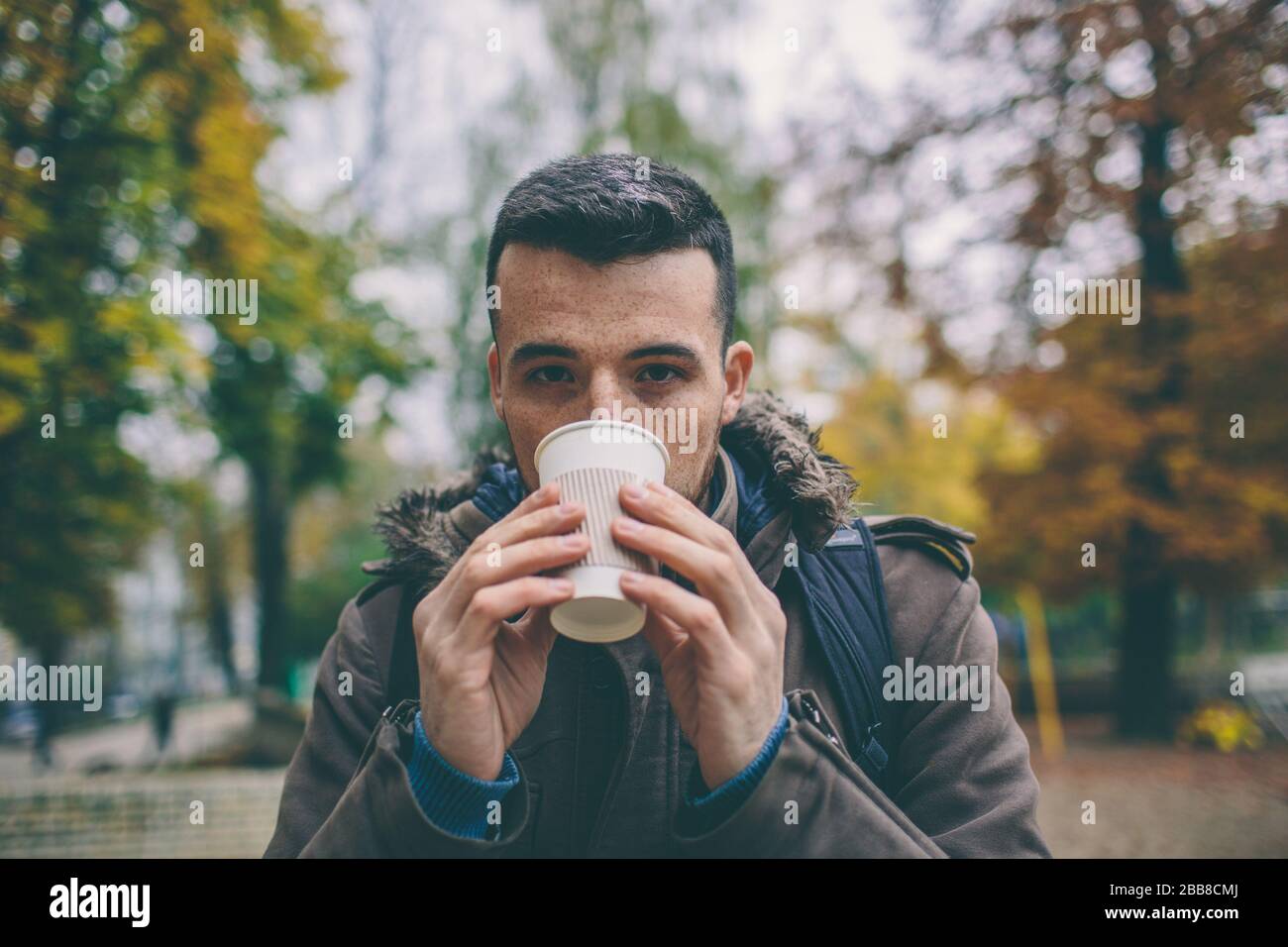 Mann im Mantel, der eine Tasse Kaffee Latte mit Milch hält. Einsame Frau steht auf einer verschneiten Herbststraße im Park. Stockfoto