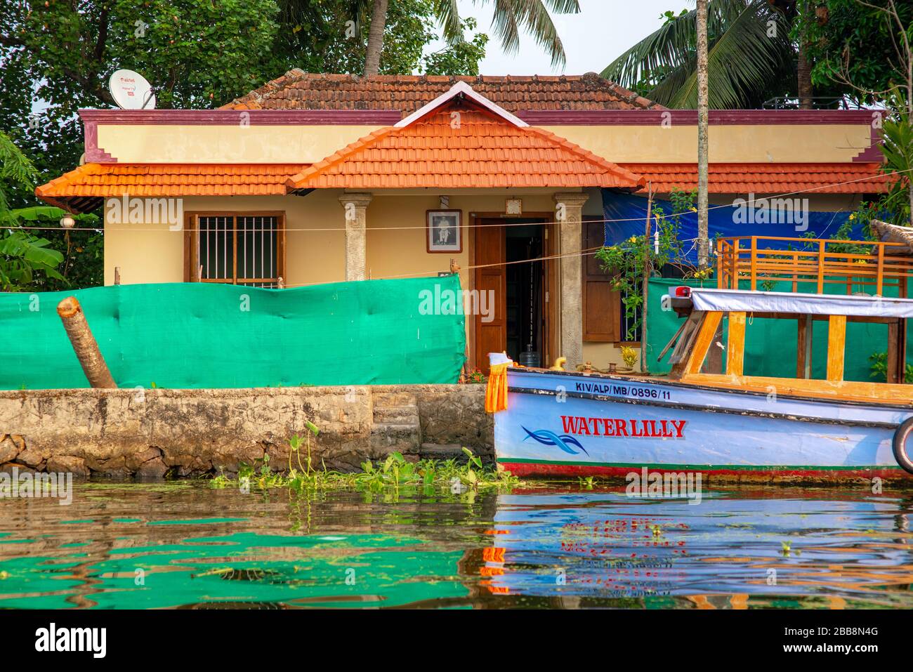 Alleppey, Kerala, Indien - 30. März 2018: Ein kleines traditionelles Haus am Rande eines Kanals und ein farbenfrohes, meist violettes Boot. Aufgenommen in Stockfoto