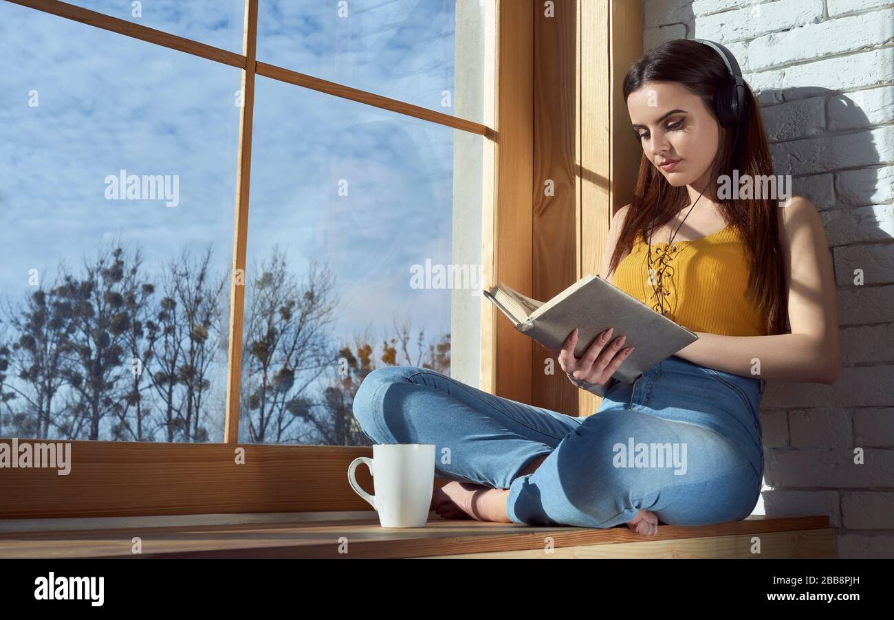 Mädchen, die auf der Fensterbank sitzen und Buch lesen, während sie Musik hören und Kaffee trinken Stockfoto