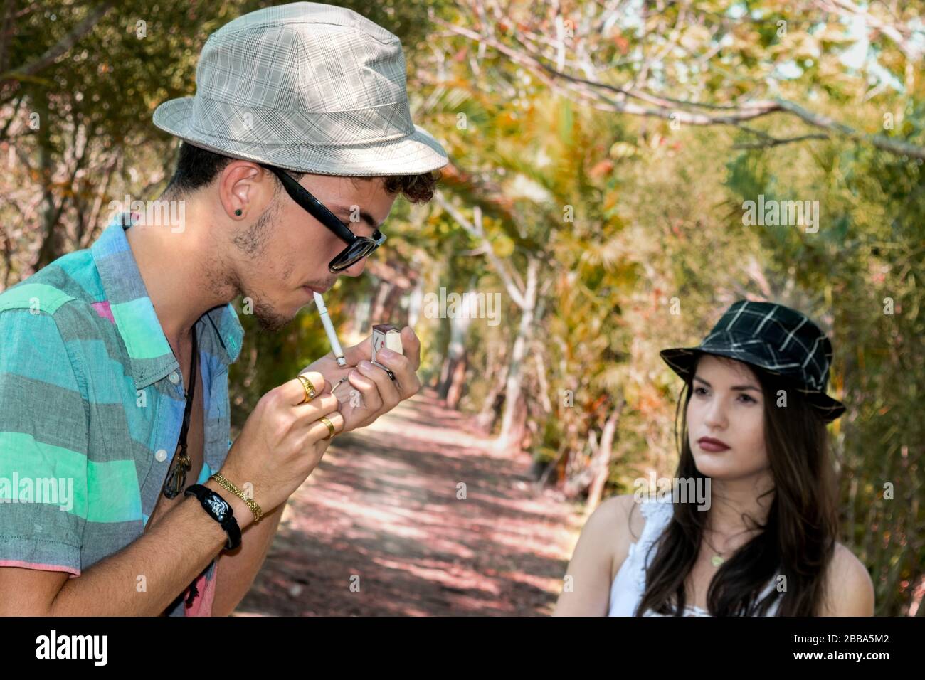 Junger kubanischer Mann, der eine Zigarette unter den Bäumen aufblitzte, während er einen missbilligenden Blick von einem jungen Mädchen in der Nähe erhielt. Stockfoto