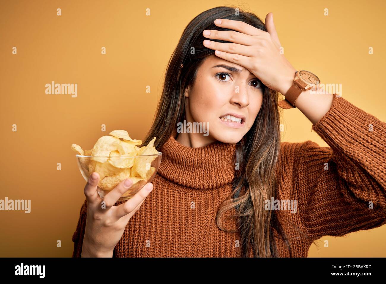 Junges, wunderschönes Mädchen, das eine Schüssel mit Kartoffelchips hält, die über gelbem Hintergrund stehen und mit der Hand auf dem Kopf gestresst ist, schockiert mit Scham und Überraschungsgesicht Stockfoto