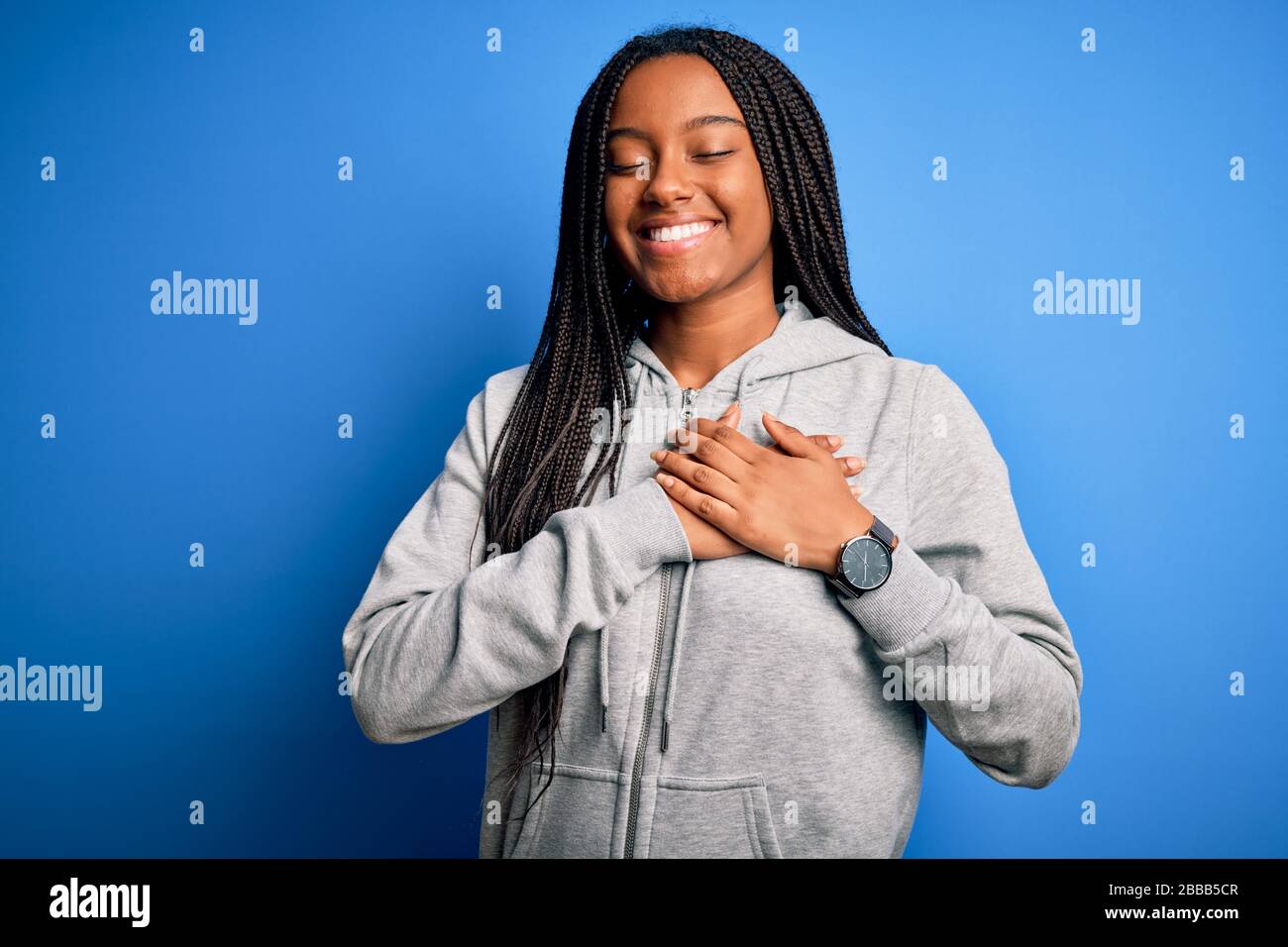 Junge afroamerikanische Sportlerin, die Sportsweatshirt über blauem isoliertem Hintergrund trägt, lächelt mit Händen auf der Brust mit geschlossenen Augen und dankbar Stockfoto