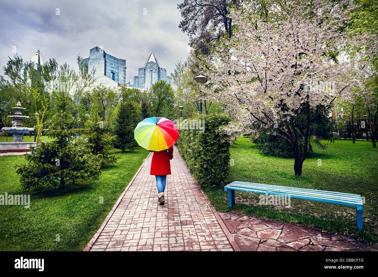 Frau im roten Mantel mit Regenbogen Regenschirm Fuß in den Park mit blühenden Bäumen bei bedecktem Himmel Stockfoto