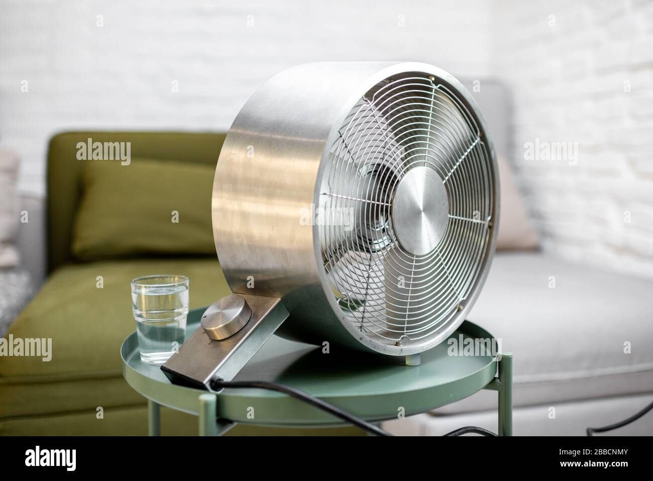 Moderner Ventilator mit einem Glas Wasser auf dem Tisch zu Hause. Konzept von anormaler Sommerhitze und Durst Stockfoto