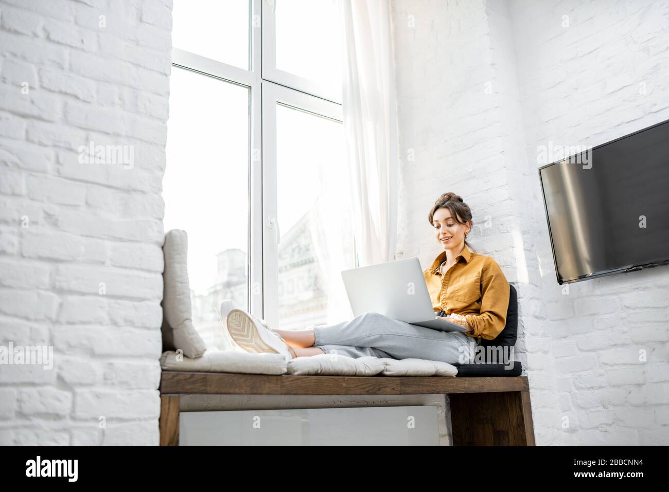 Junge Frau kleidete sich beiläufig am Laptop, während sie zu Hause auf dem Fensterschweller saß. Arbeiten Sie von zu Hause aus in gemütlichem Atmospha-Konzept Stockfoto