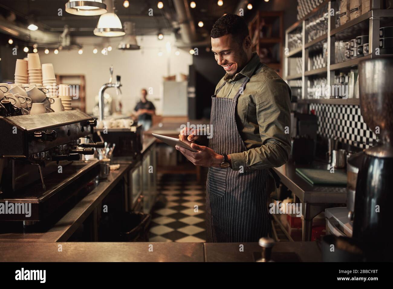 Fröhlicher junger afro-amerikanischer Café-Besitzer, der schwarze gestreifte Schürze mit digitalem Tablet trägt Stockfoto