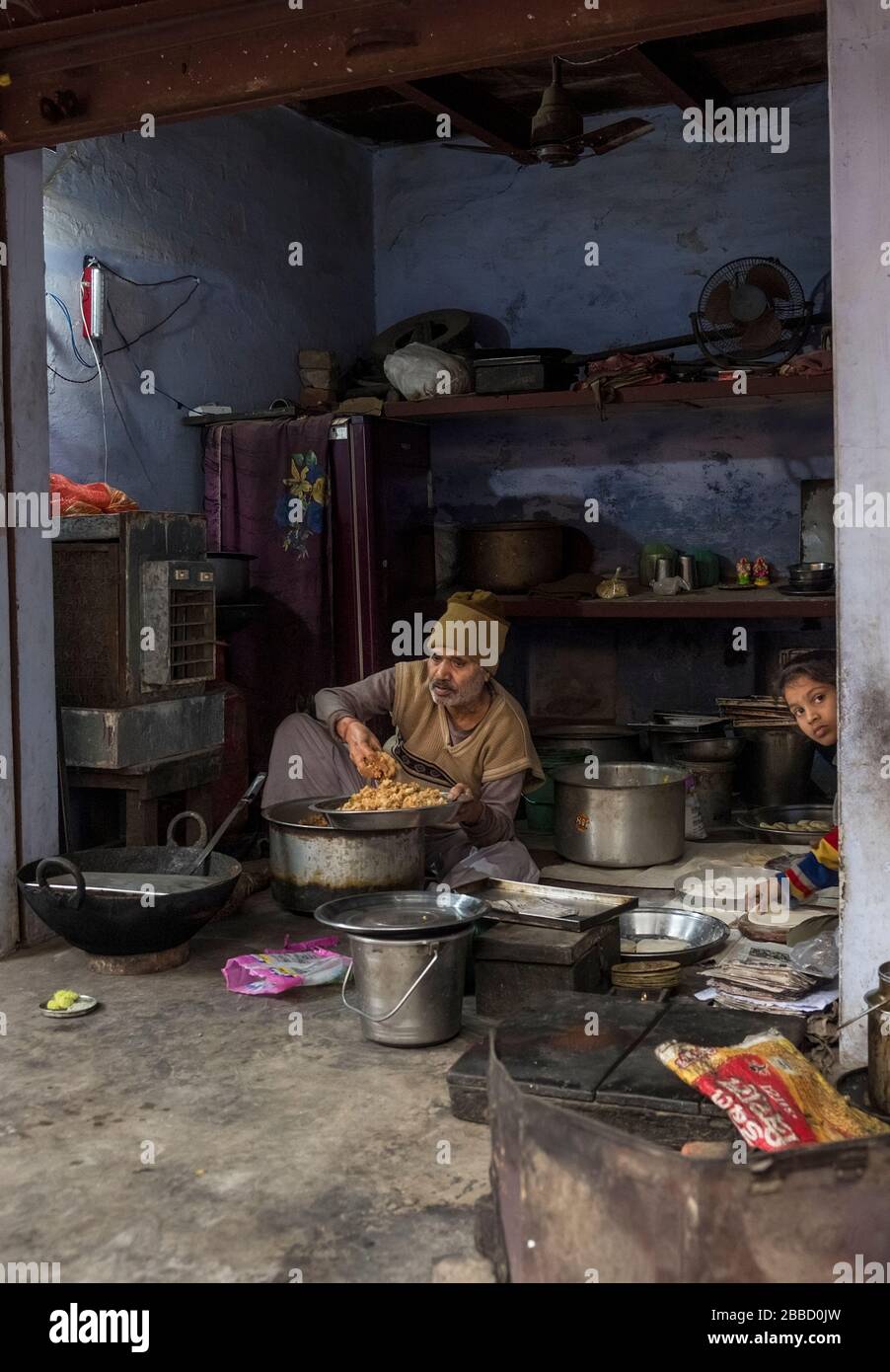 Ein Inder hockt auf dem Boden einer kleinen, vollgestopften Küche, umgeben von Töpfen Stockfoto
