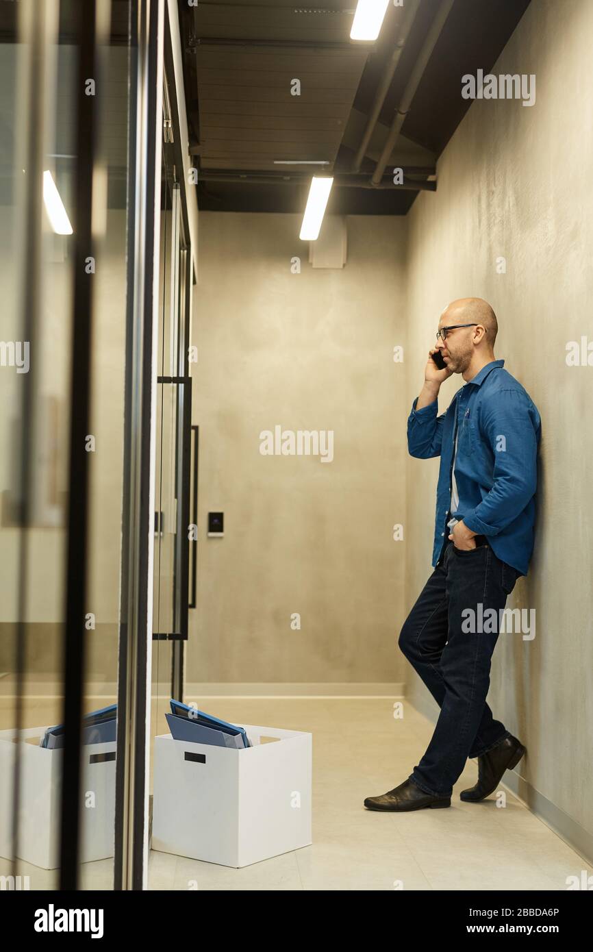 Seitenansicht in voller Länge bei einem kahlen erwachsenen Mann, der mit dem Smartphone spricht, während er sich an die Wand in einer minimalen Bürohalle, einem Kopierraum, lehnt Stockfoto