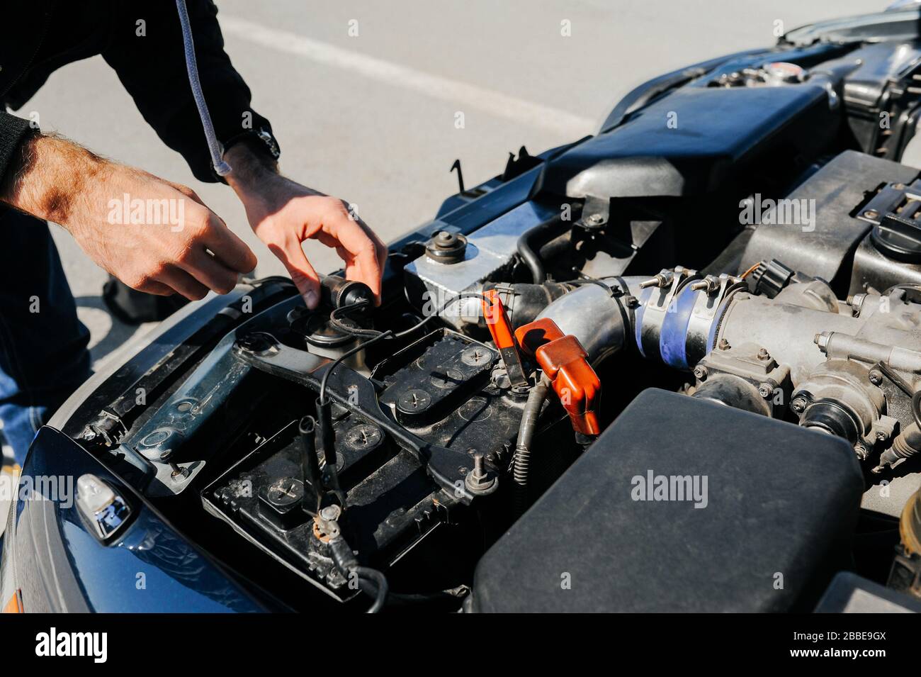 Die Hand verbindet die rote Klemme der Zange mit der Batterie eines Autos mit offener Haube und abgestimmten Teilen des Wagens Stockfoto