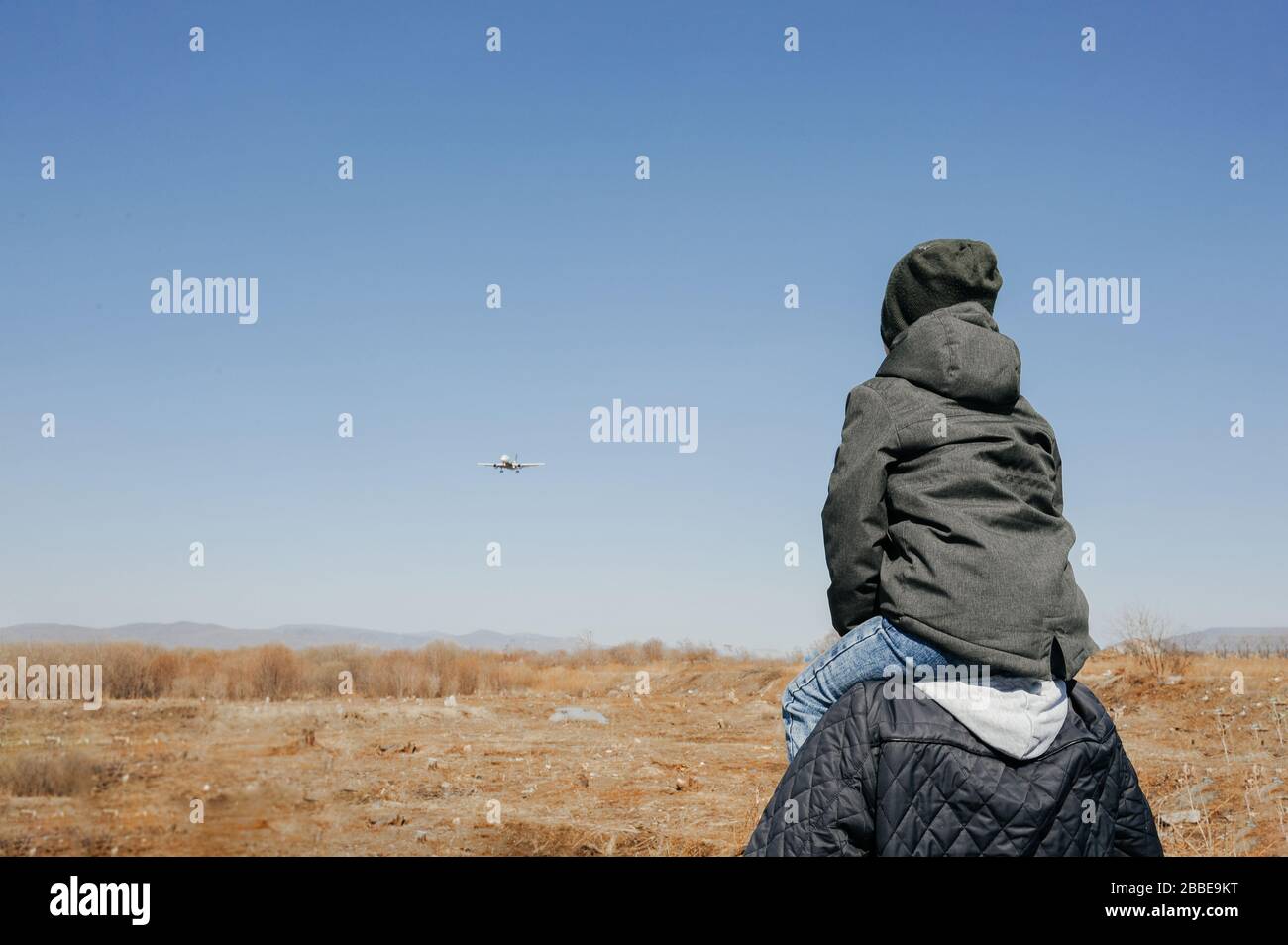 Ein Junge in einer Khaki-Jacke und einem Hut sitzt auf den Schultern eines gewachsenen Mannes und sie beobachten zusammen, wie ein Passagierflugzeug in einem klaren blauen Himmel fliegt Stockfoto