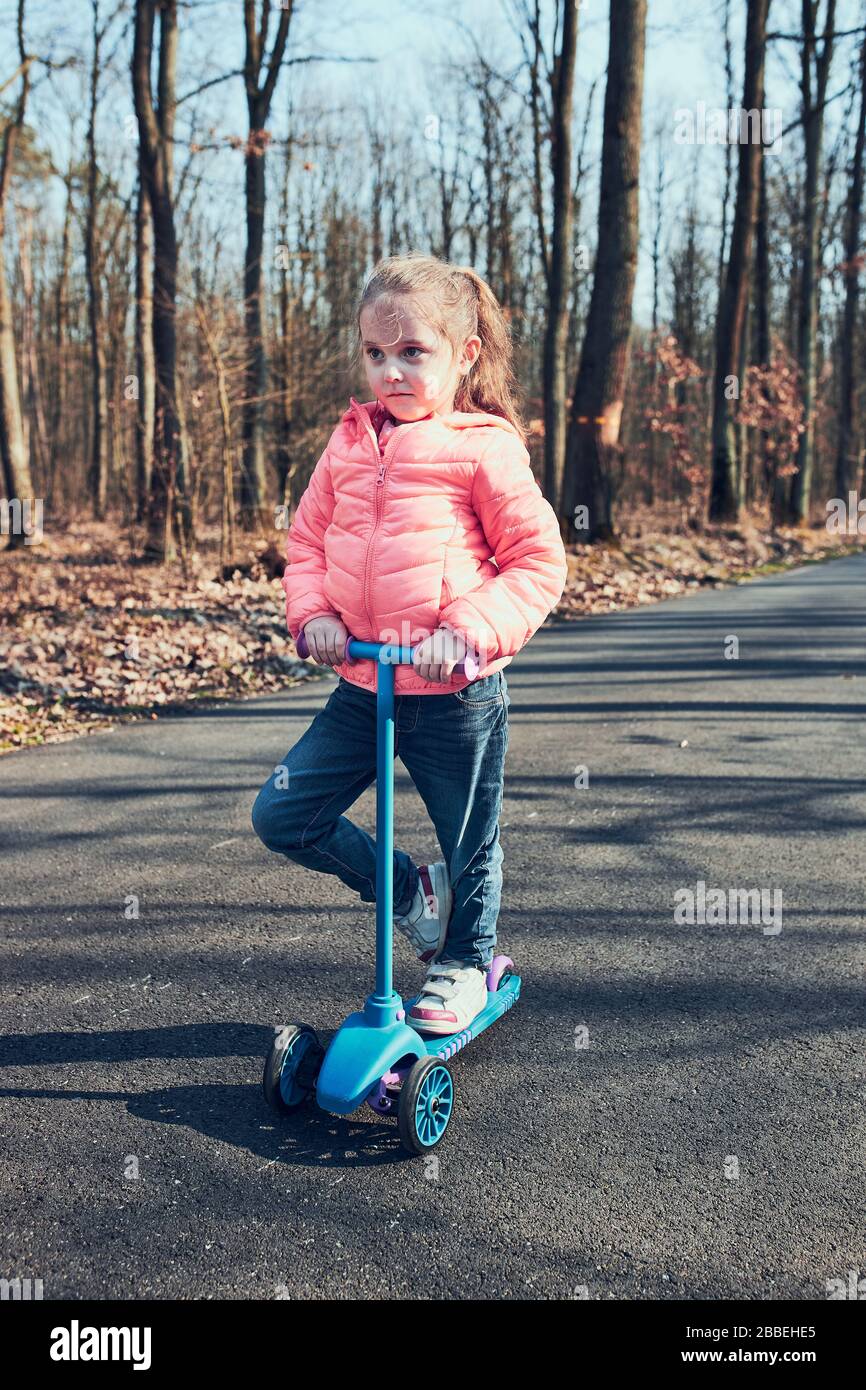 Kleines Mädchen, das im Park spielt, einen Roller fährt und Spaß am sonnigen Herbsttag hat. Echte Menschen, authentische Situationen Stockfoto