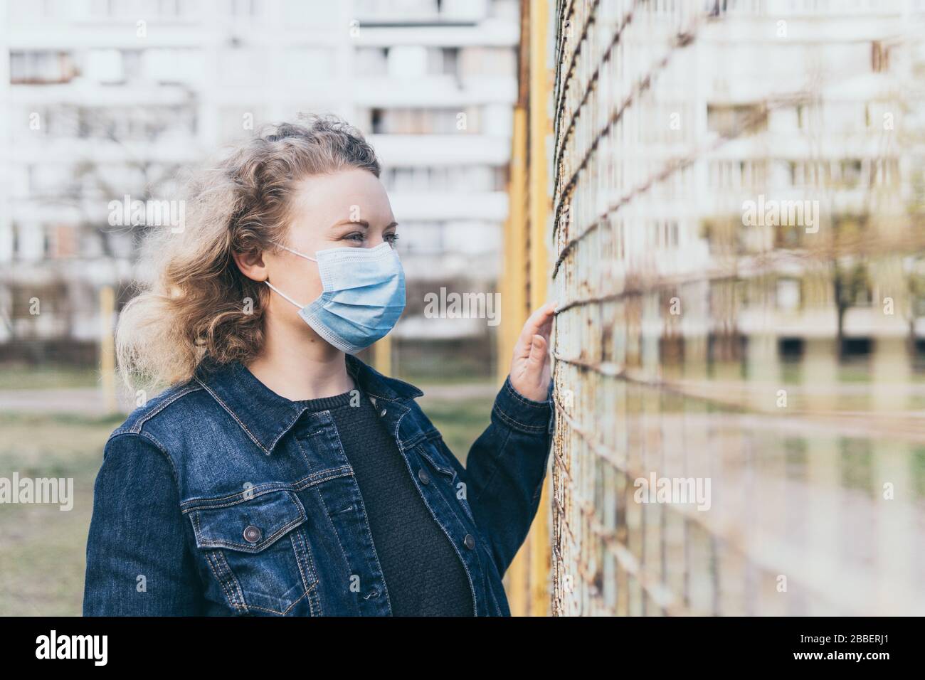Osteuropäerin besorgt Frau, die im Freien medizinische Maske trägt, neben dem Gitterzaun mit Wohnhaus auf dem Hintergrund steht. Corona-Virus Stockfoto