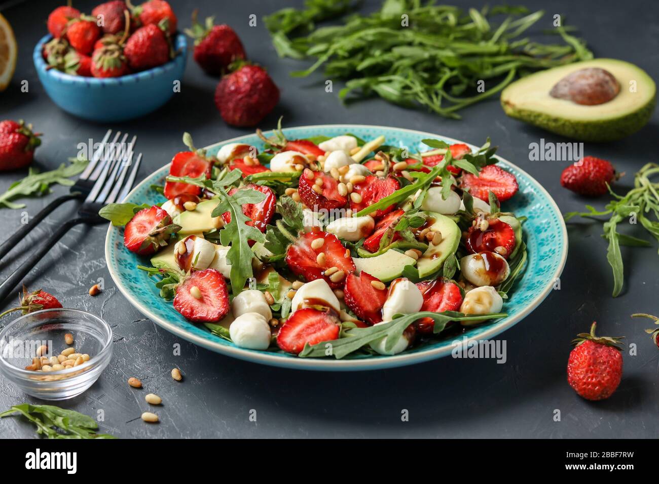 Gesunder Salat mit Erdbeeren, Avocado, Arugula und Mozzarella, gekleidet mit Olivenöl und Balsamico-Dressing in einem blauen Teller auf dunklem Hintergrund Stockfoto