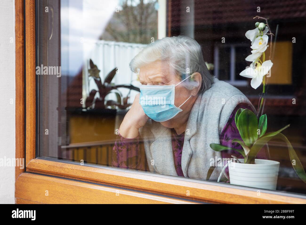Einsame Seniorin mit chirurgischer Maske, die zu Hause auf einer Fensterebene sitzt, Coronavirus und Kovid-19-Bestimmungen Stockfoto