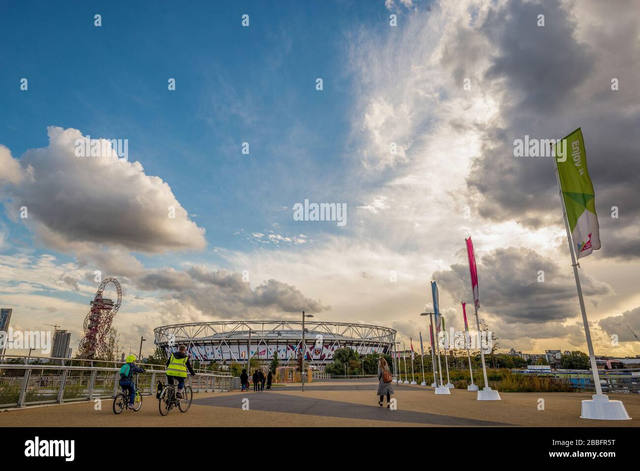 Die Sonne geht über das Olympiastadion 2012, in dem jetzt das Fußballteam West Ham United beheimatet ist. Die Menschen gehen und radeln auf dem Weg, der zum Boden führt. Stockfoto