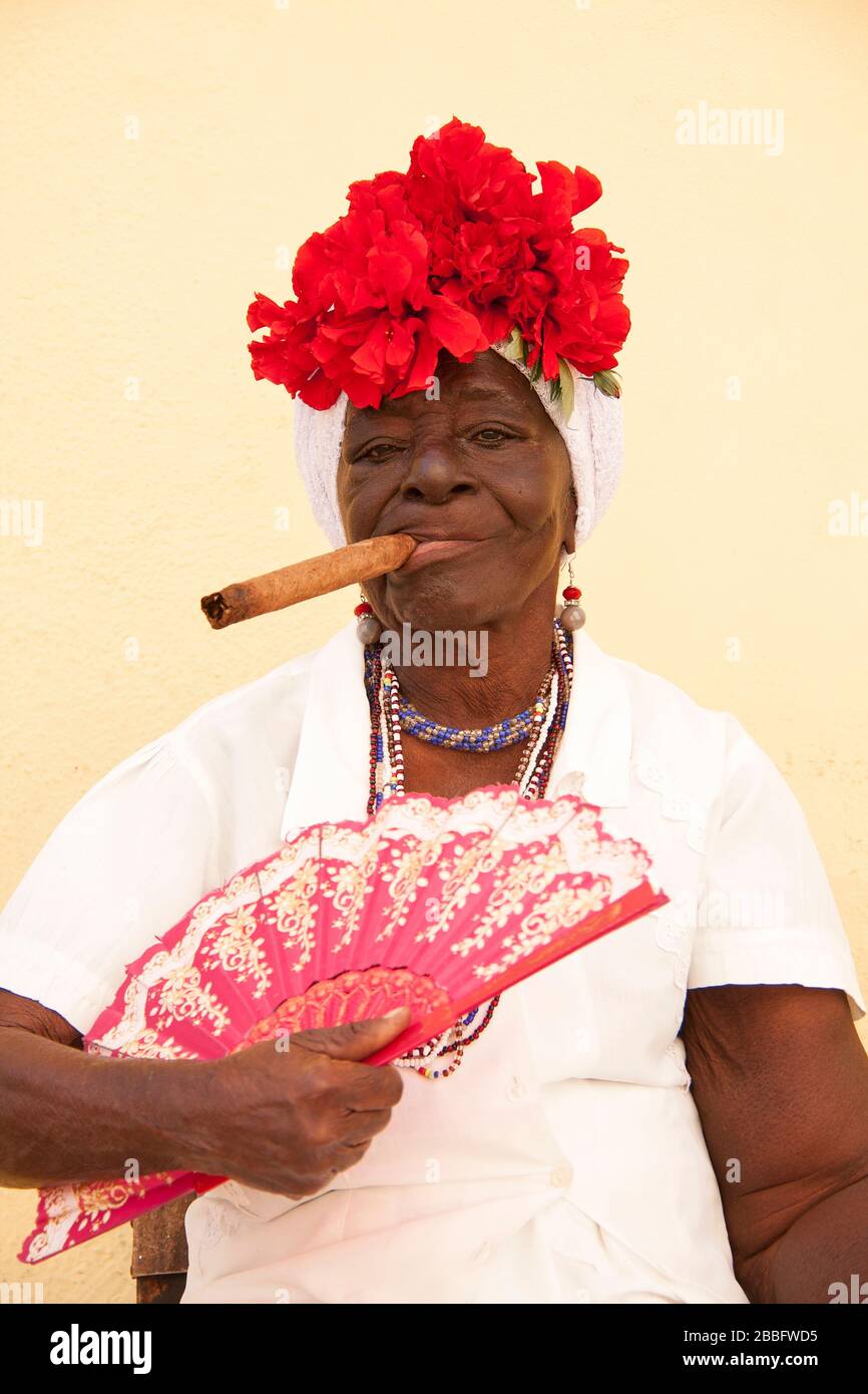 HAVANNA, KUBA - 29. MÄRZ 2017: Porträt einer alten schwarzen Frau in typischer Havannaer Kleidung, die eine große kubanische Zigarre an der pastellgelben Wand raucht Stockfoto