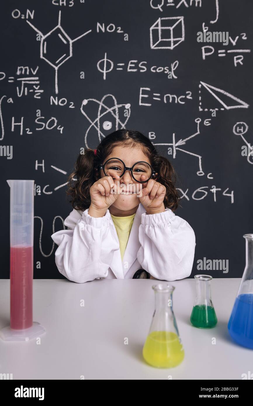 Lustige kleine Wissenschaftlerin im Laborkittel scherzt mit ihrer Brille, auf dem schwarzen Brett mit wissenschaftlichen Formeln, dem Konzept der Rückkehr zur Schule und dem Erfolg Stockfoto