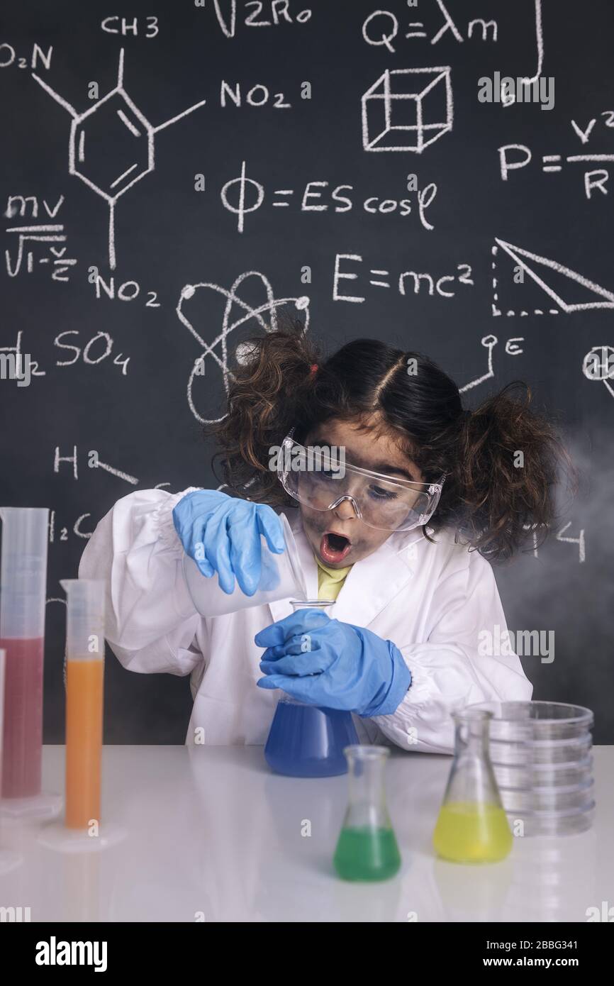 Überraschtes Mädchen der Wissenschaftlerin mit Handschuhen und Brille im Labormantel, das chemische Kolben mischt, Hintergrund der Tafel mit wissenschaftlichen Formeln, Explosion im Labor Stockfoto