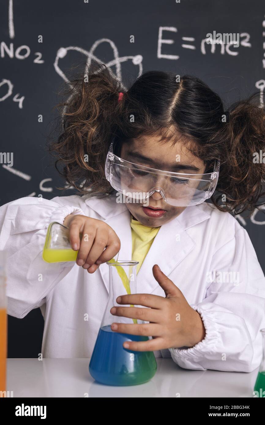 Wissenschaftler Kind mit Brille und Handschuhen im Labormantel, das chemische Flüssigkeiten in Kolben mischt, Hintergrund auf der Tafel mit wissenschaftlichen Formeln, Explosion im Labor Stockfoto