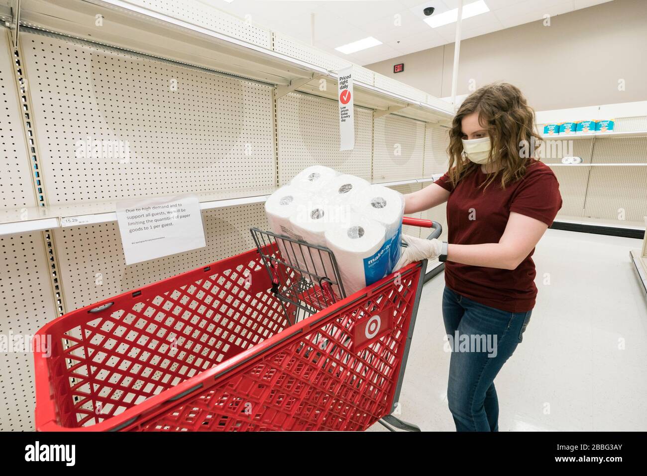 Indiana, USA - 24. März 2020: Eine blonde Frau, die eine Gesichtsmaske und Handschuhe trägt, legt ein Paket von Papiertüchern in ihren Warenkorb inmitten leerer Regale Stockfoto