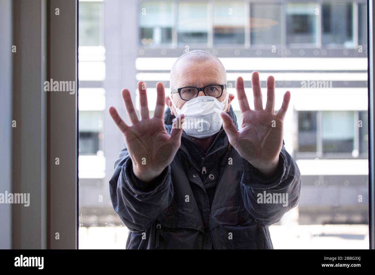 Mann mit medizinischer Gesichtsmaske, der außerhalb eines Fensterabschnitts steht, wobei seine Hände gegen Fensterglas gedrückt und nach innen schauen. Corona Covid-19 Virus qua Stockfoto