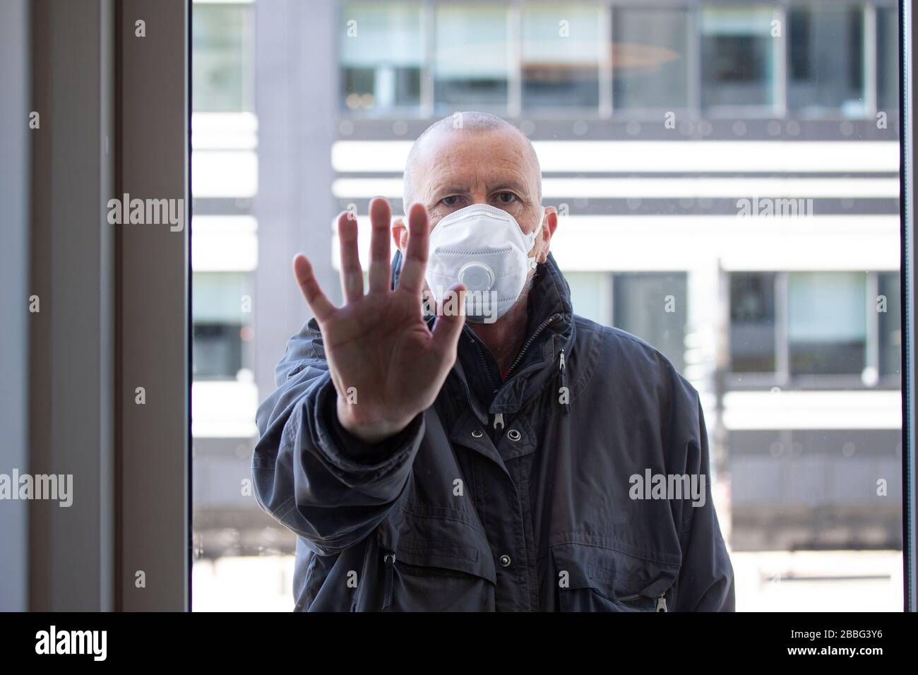 Mann mit medizinischer Gesichtsmaske, der außerhalb eines Fensterabschnitts steht, wobei die rechte Hand gegen das Fensterglas gedrückt und nach innen blickt. Corona Covid-19 Viru Stockfoto