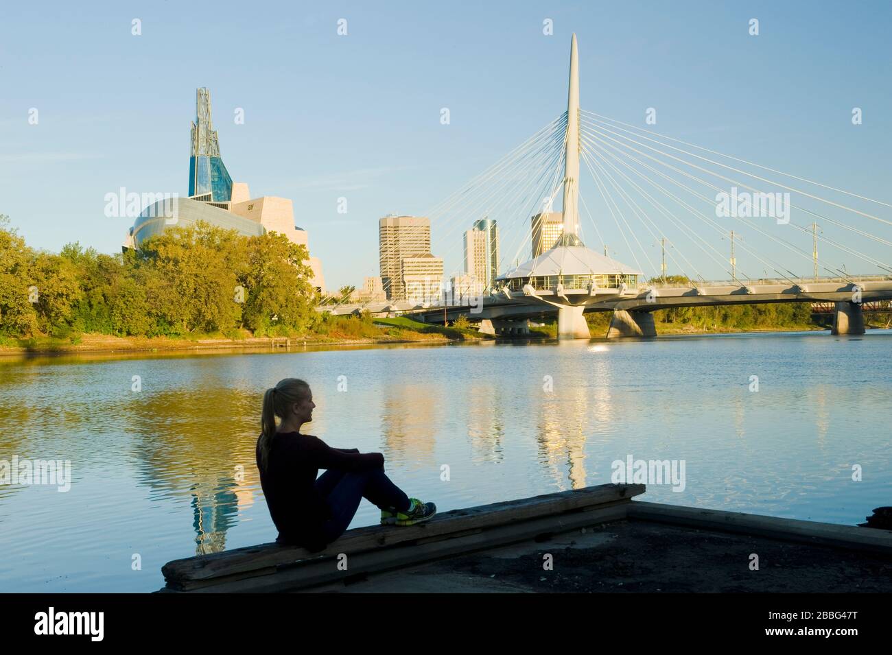 Mädchen entspannen, Skyline von Winnipeg von St. Bonifatius mit Blick auf den Red River, die Esplanade Riel Bridge und das Canadian Museum for Human Rights, Manitoba, Kanada Stockfoto