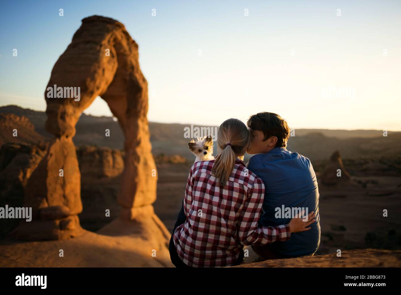 Junger Mann küsst seine Freundin auf die Wange, während ihr Hund über ihre Schulter blickt und im Hintergrund eine malerische Schlucht sieht Stockfoto