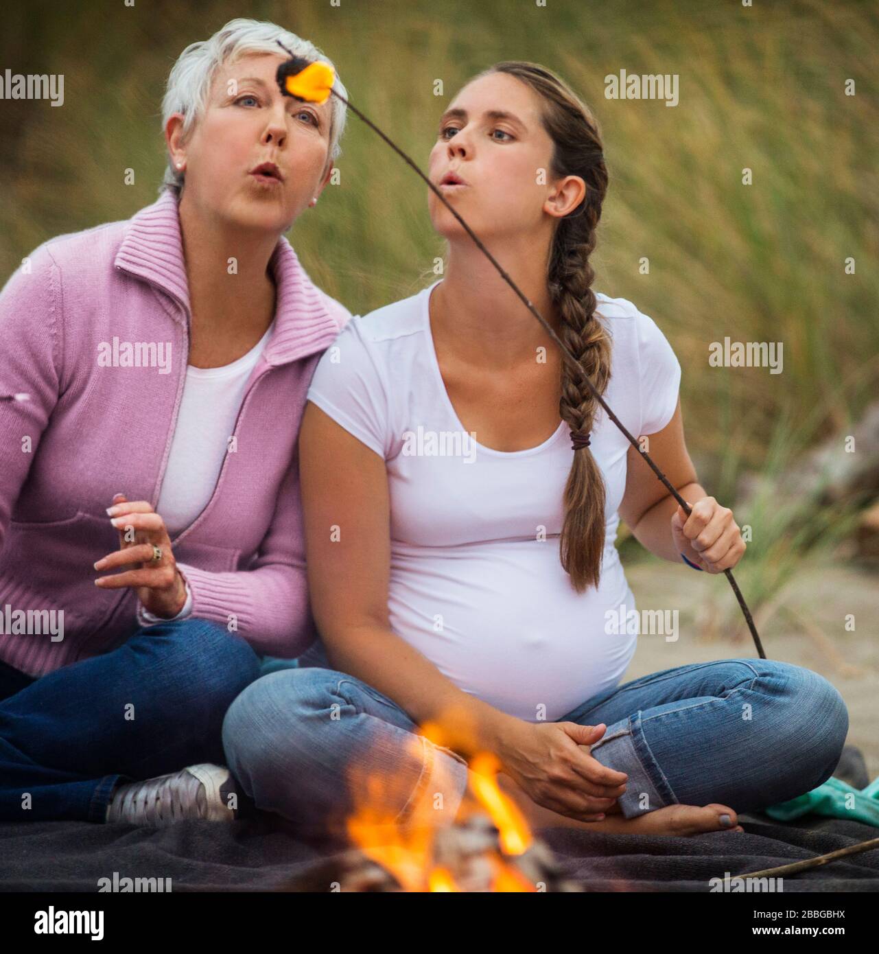 Die reife Frau und ihre schwangere Tochter bläsen während eines Lagerfeuers am Strand auf einen brennenden Marshmallow Stockfoto