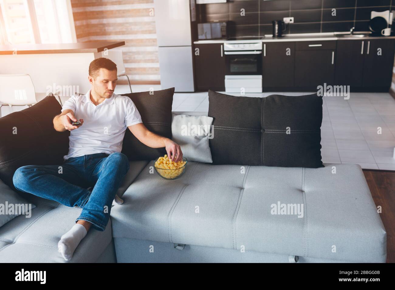 Der junge Mann gucken in seiner eigenen Wohnung fernsehen. Der konzentrierte Kerl hält die Fernbedienung in der Hand und bekommt einen Snack aus der Schüssel. Sehen Sie sich Filme oder fernsehsendungen allein im Zimmer an Stockfoto