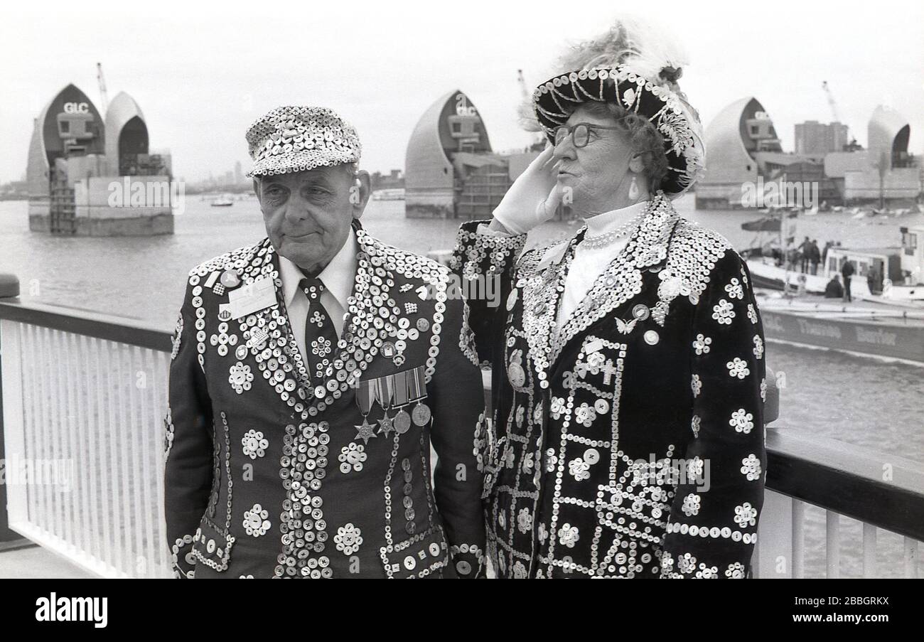 1984 tragen Londons Perlkönig und Königin ihre traditionellen Outfits. Jacken mit Perlmuttknöpfen, die zusammen an den Royal Docks stehen, bei der Eröffnung der neuen Thames Barrier, South London, England, Großbritannien durch Königin Elizabeth II..stellvertretend für Londons Arbeiterkultur, "Pearlies", wie sie auch bekannt sind, Entstanden aus den Coster Kings und Queens, die als Führer der Londoner Straßenhändler gewählt wurden. Stockfoto