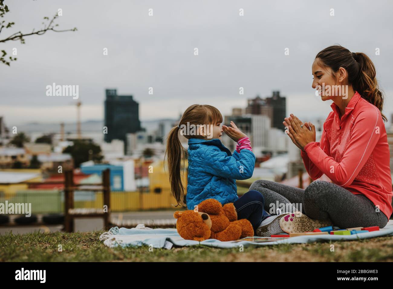 Seitenansicht einer fröhlichen lächelnden Frau und eines Mädchen-Kindes, das beim Sitzen in einem Park Patty-Kuchen-Spiel spielt. Kleines Mädchen spielt Klatschspiel mit ihrem Kindermädchen ou Stockfoto