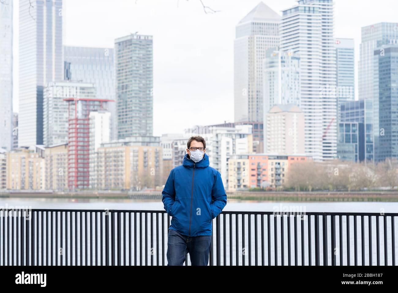 Männer aus Mitte 40 tragen eine blaue Jacke und eine Gesichtsmaske, die auf der Themse entlang spazieren, mit Londons Finanzbezirk und Canary Wharf im Hintergrund. Stockfoto