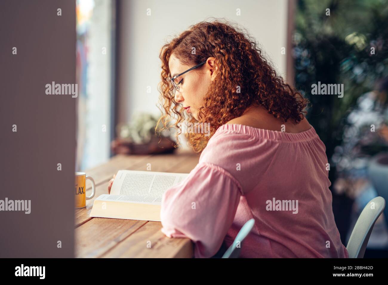 Junge rote Haarfrau mit Brille sitzt und liest ein Buch. Stockfoto