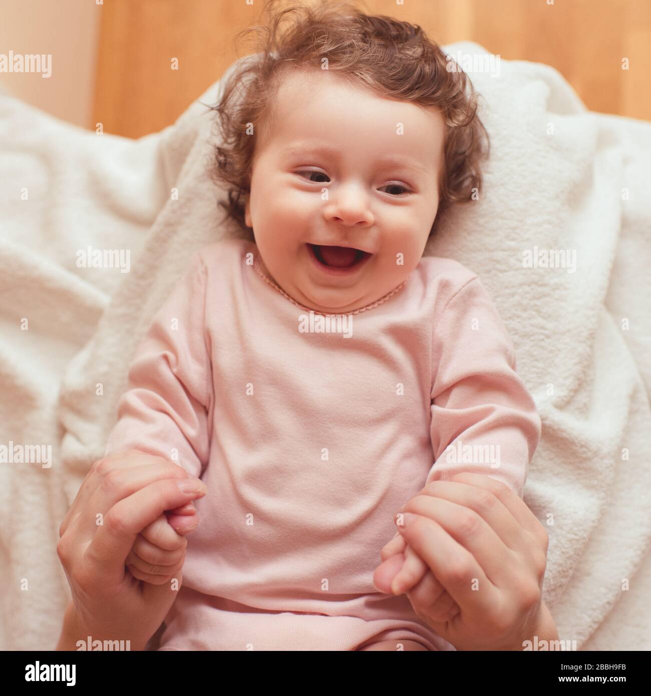 Glückliches Baby mit lockigen Haaren, die im Bett liegen und die Hände der Mutter umhüllen. Draufsicht. Mutterschaft. Mutterschaft. Kindheit. Stockfoto