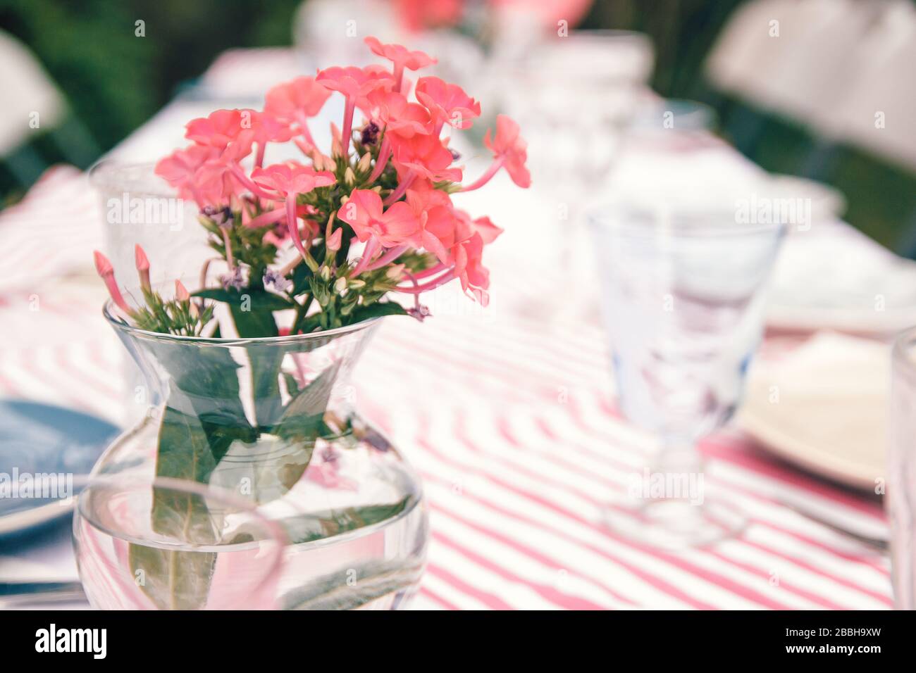 Ländlicher Hochzeitstisch mit Korallenblumen in einer Glasvase auf einem gestreiften Tischtuch. Spezielle Ereignistabelle eingerichtet. Frische Blumendekoration. Stockfoto