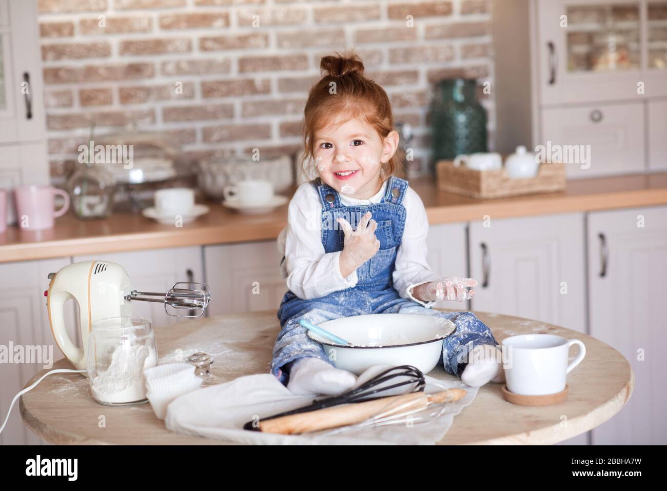 Das lachende Mädchen 3-4 Jahre alt sitzt auf dem Tisch in der Küche und macht Kuchen mit Teig Closeup. Blick auf die Kamera. Kindheit. Stockfoto
